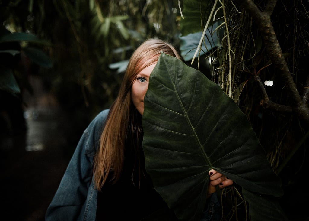 White female hidden among tropical plants. She is holding a large leaf to obscure her fact, but you can still see her long brown hair and blue top.
