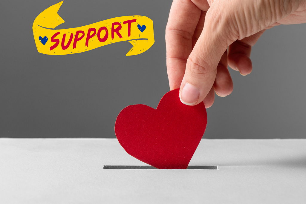 A hand inserting a heart-shaped paper cutout into a donation box, with a banner labeled ‘Support’ next to the hand.