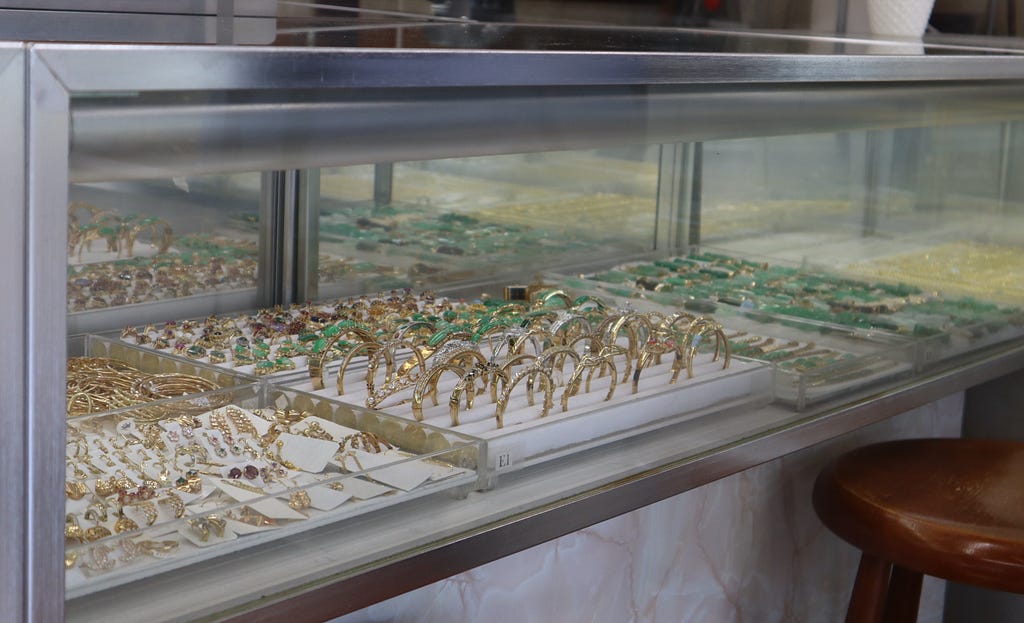 Gold and jade jewelry are displayed inside one of the many glass cases in the shop — bracelets, rings, earrings, and necklaces of all shapes, sizes, and variations. A chestnut brown stool stands at the bottom right corner, mostly out of frame.