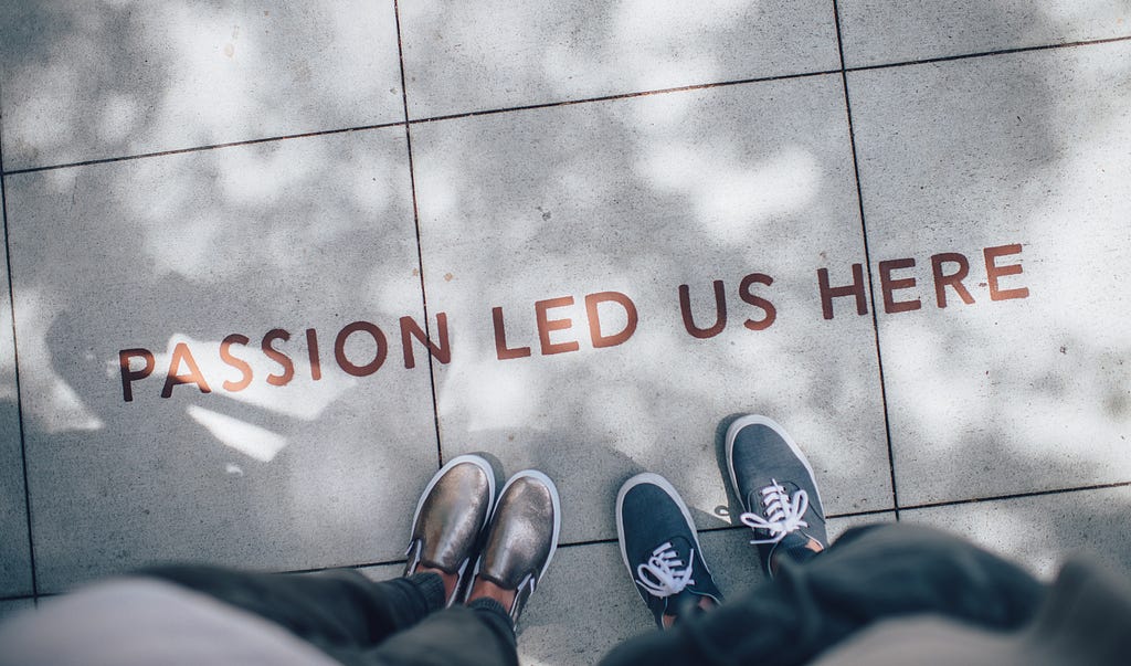 The sign “Passion led us here” on the sidewalk, two people, from the perspective of the standing person