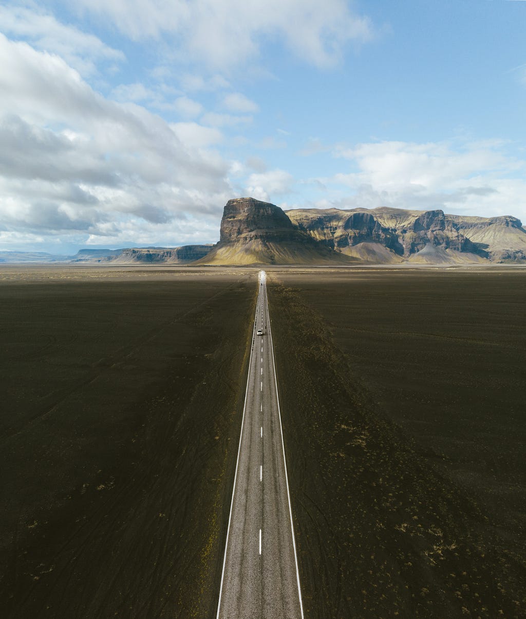 Aerial view of a long straight stretch of road leading toward desert plateaus. A single car is driving on the road, implying a long journey in a vast land of possibility.