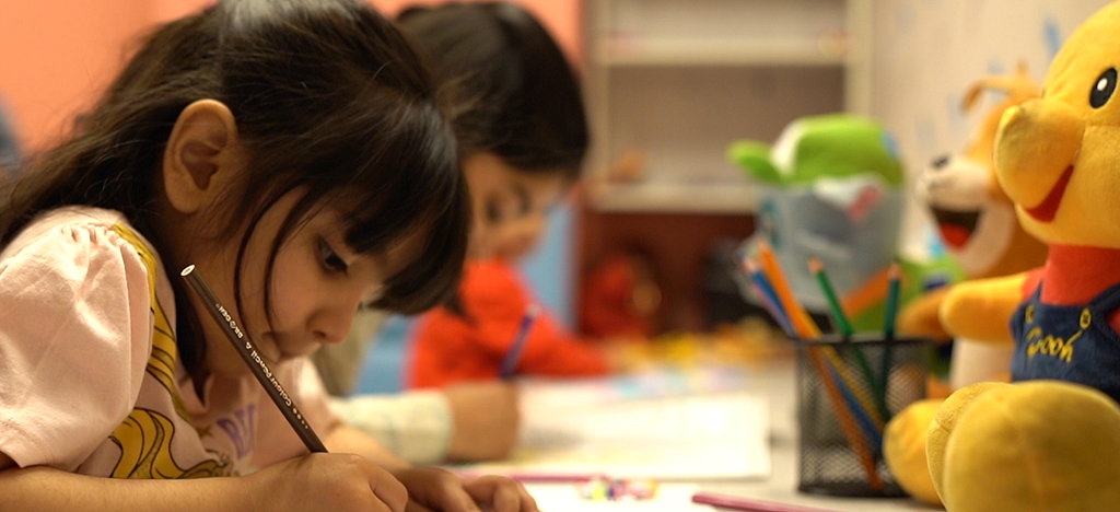 A little girl writing on a piece of paper, in a playroom at Daftarkhwan | Vogue.