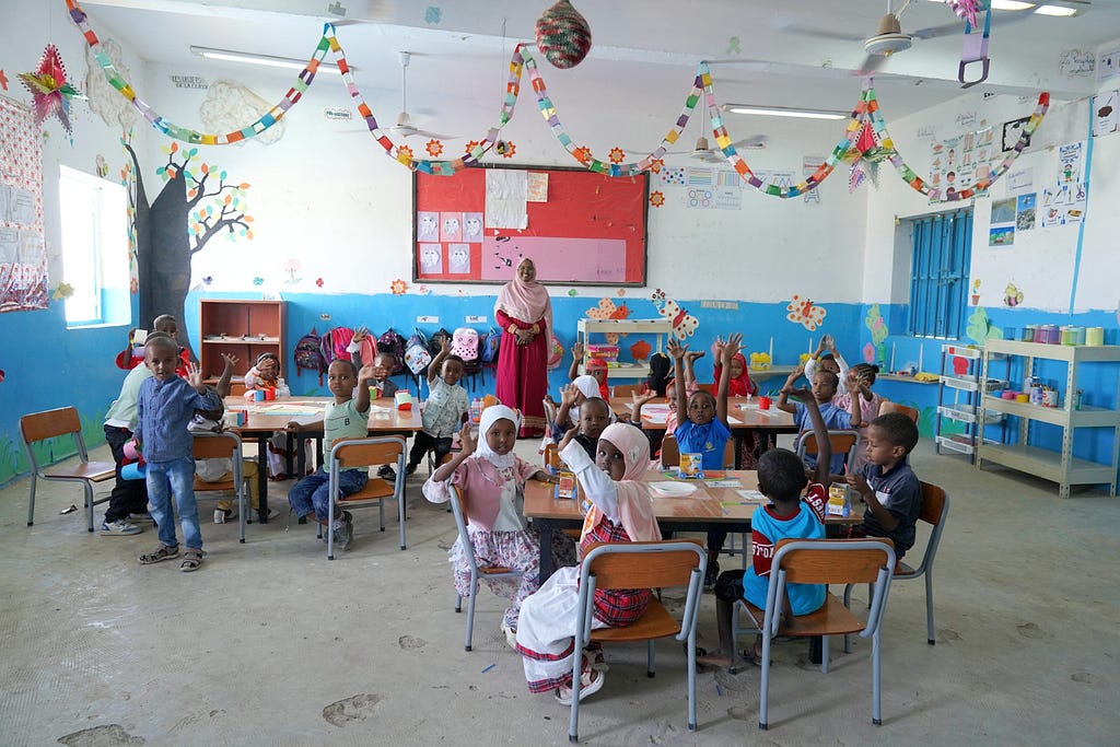 A classroom of preschool students raise their hands in the air as their teacher stands at the head of the classroom.