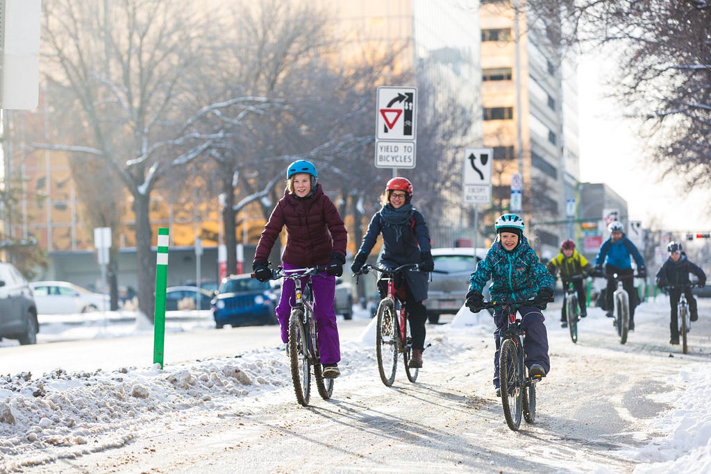 Kids and adults in full snow gear ride their bikes down a plowed bike lane in the winter.