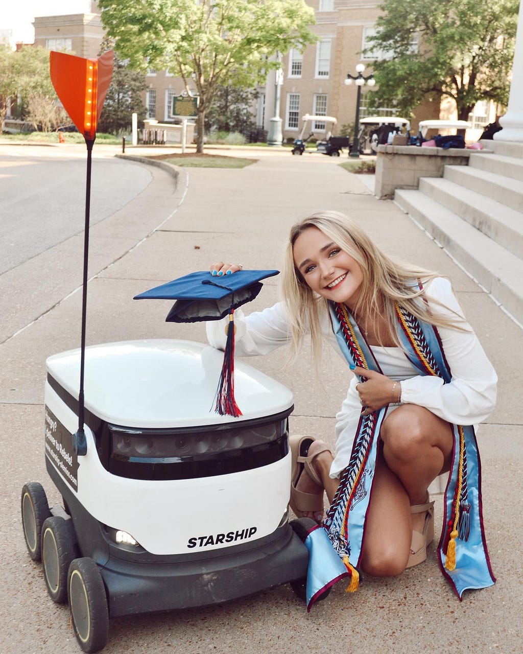 Mercy posing with a Starship Delivery Robot, holding her blue graduation cap over the robot’s lid.