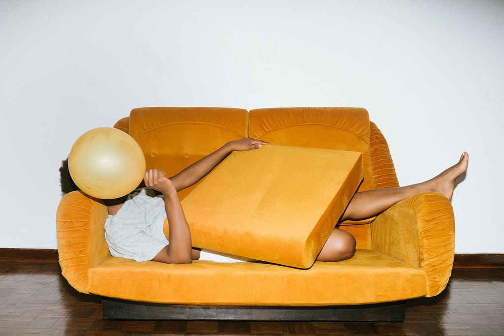 A person stretched out on a mustard colored couch with a balloon covering the face