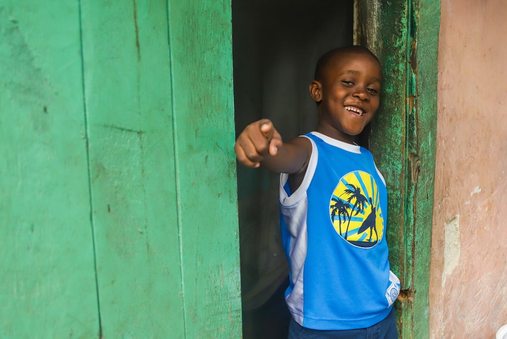 A Haitian boy stands in a green doorway wearing a blue t-shirt and smiling and posing, pointing his finger at the camera.