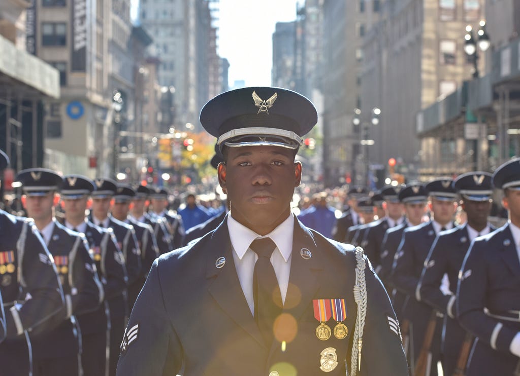 A young man with dark brown skin wears a United States Air Force uniform and stands in the streets of New York City flanked by fellow airmen for the Veterans Day Parade in 2018.