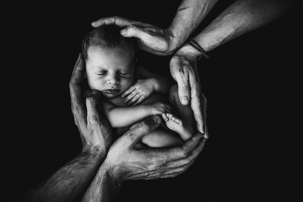 Two sets of wrinkled hands cradle a small baby from either side. Only the baby and the hands are visible. The background is totally black.