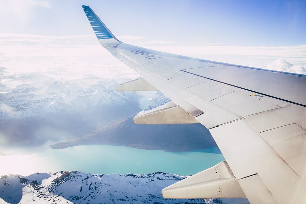 Asa de avião sob um céu azul com montanhas nevadas e um lago verde.