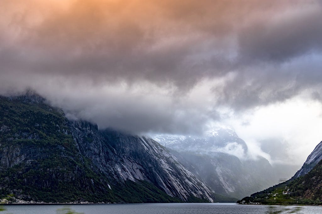 A picturesque fjord. Snow speckles the rough mountainsides. Thick clouds with a orange hue in the top left from sunlight.