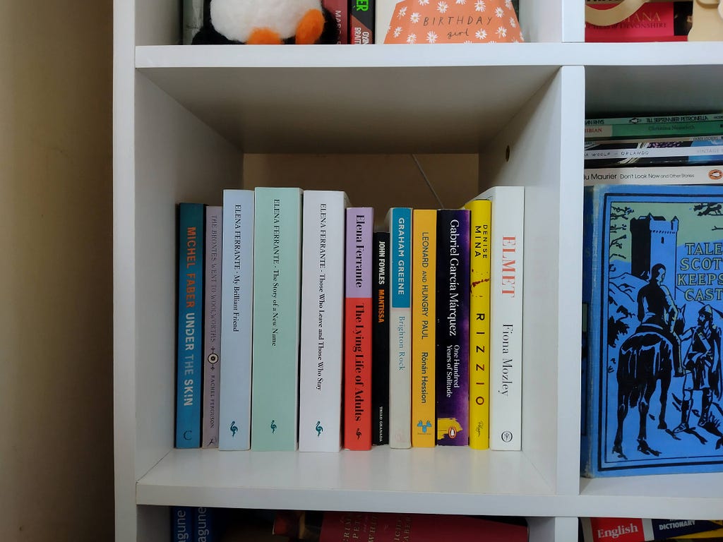 A bookshelf of colourful books. Several are written by Elena Ferrante.