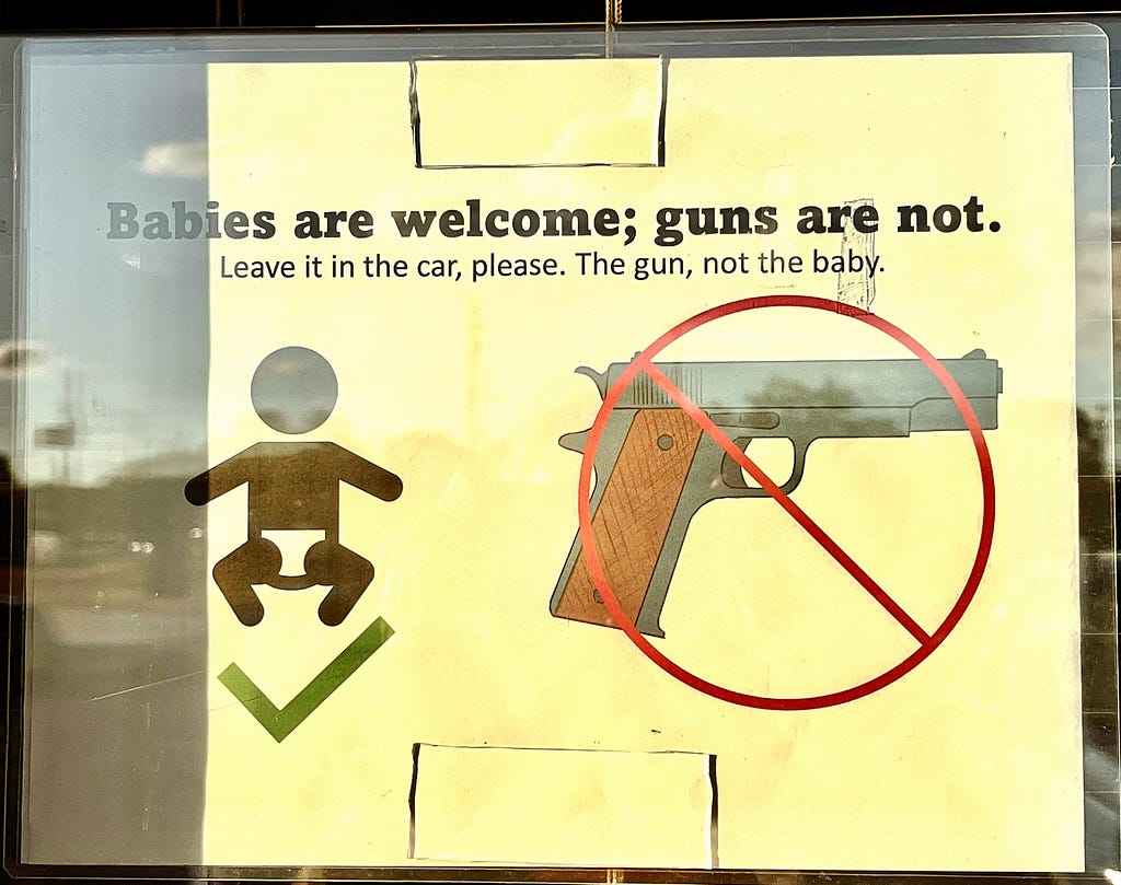 Color closeup photo of a yellowed (from the sun) sign on a glass store window showing a black graphic figure of a baby with a ‘checkmark’ underneath it and to the right beside it a black graphic of a gun pointing to the right with a red circle and line crossed over the gun (as in ‘no’), with the black text statement above both stating: “Babies are welcome; guns are not. Leave it in the car, please. The gun, not the baby.”