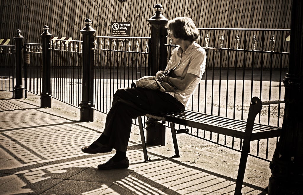 An older woman sits on a bench in front of a fence in the sun. Sepia coloured image.