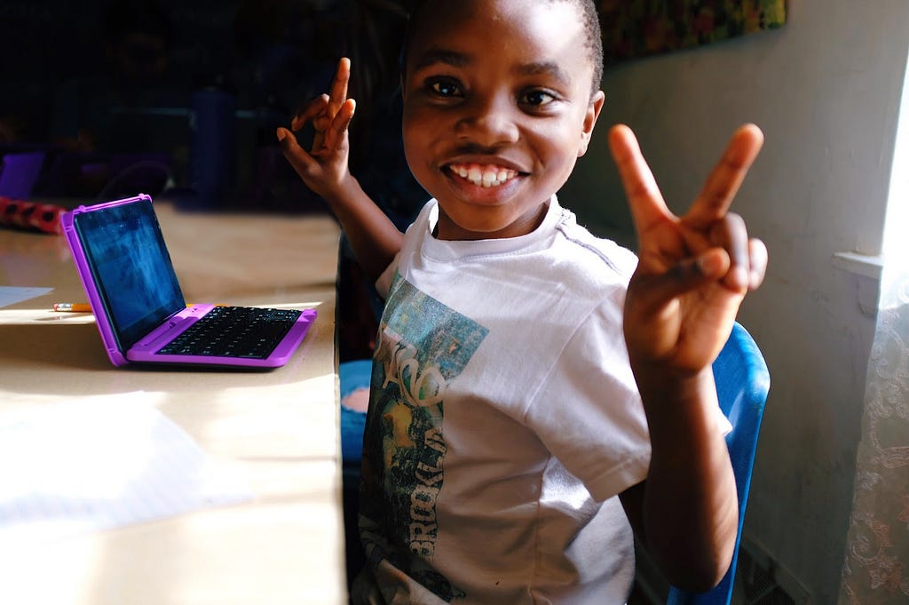 Image of a boy sitting at a desk at school, looking at the camera and making the V for Victory with both his hands. He has a tablet in front of him and looks happy.