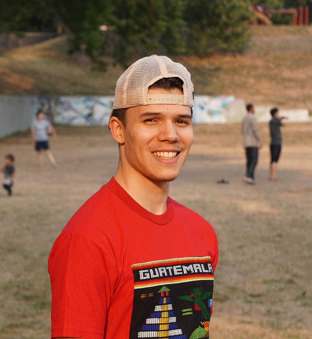 Peter, a light-skinned man with short dark hair, stands on a beach, wearing a red shirt that says Guatemala above a traditional design and a backwards, snap-back trucker hat. He is looking at the camera and smiling.