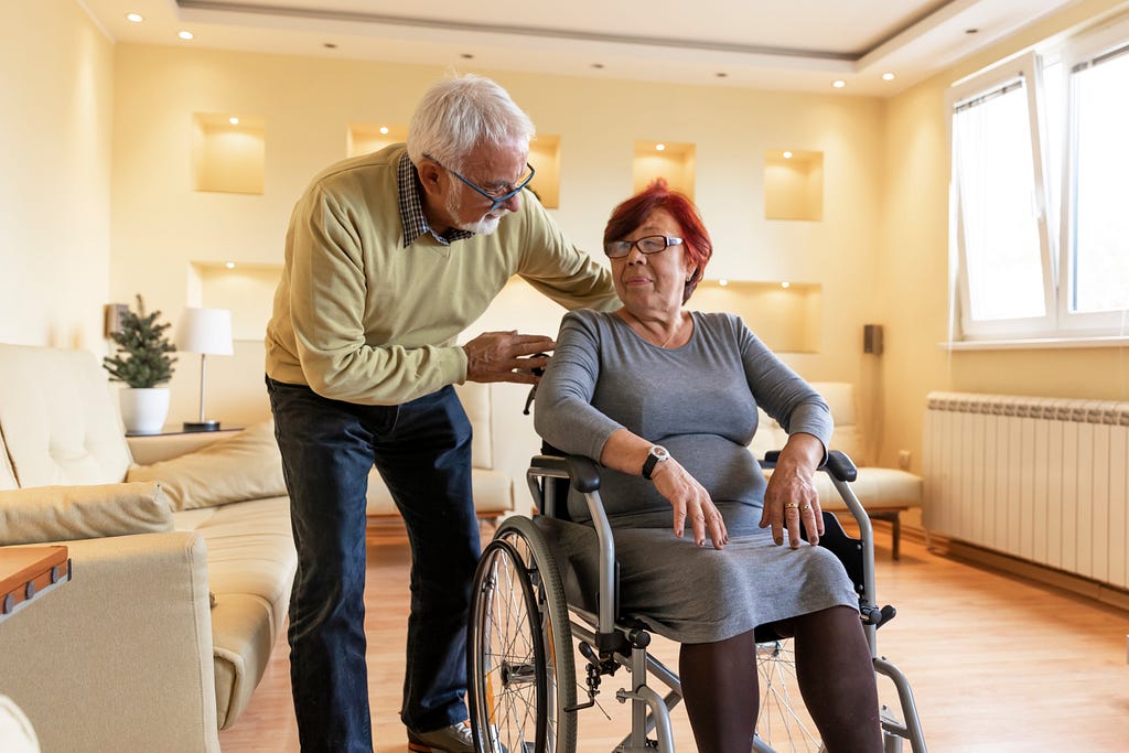 Older Asian woman in wheelchair looking at an older White man leaning over her shoulder to talk to her. Photo by ProfessionalStudioImages/Getty Images