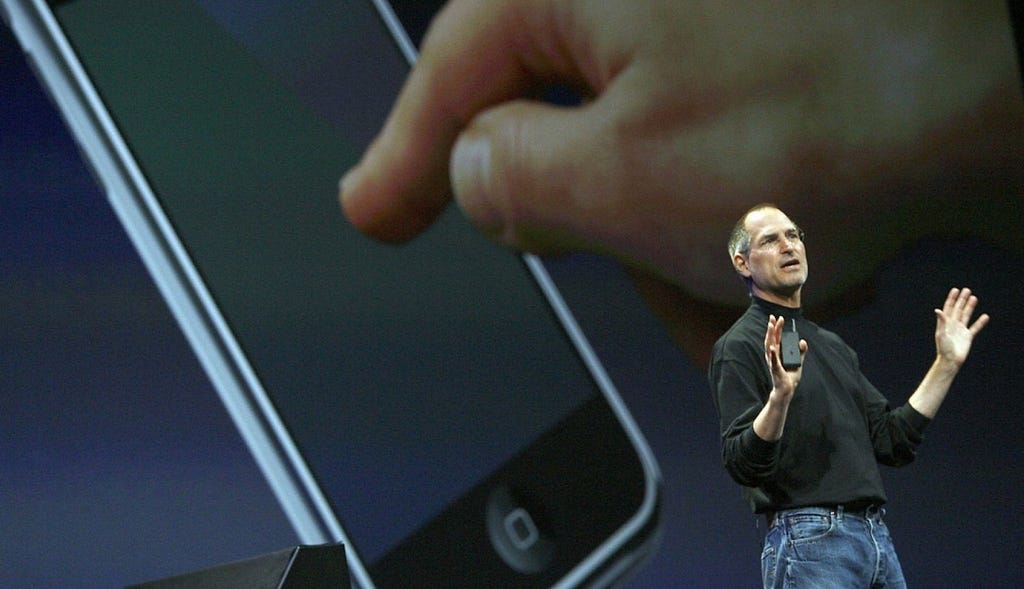 A photo from the original iPhone announcement. A large screen shows an image of someone touching an iPhone with their finger, while Steve Jobs presents