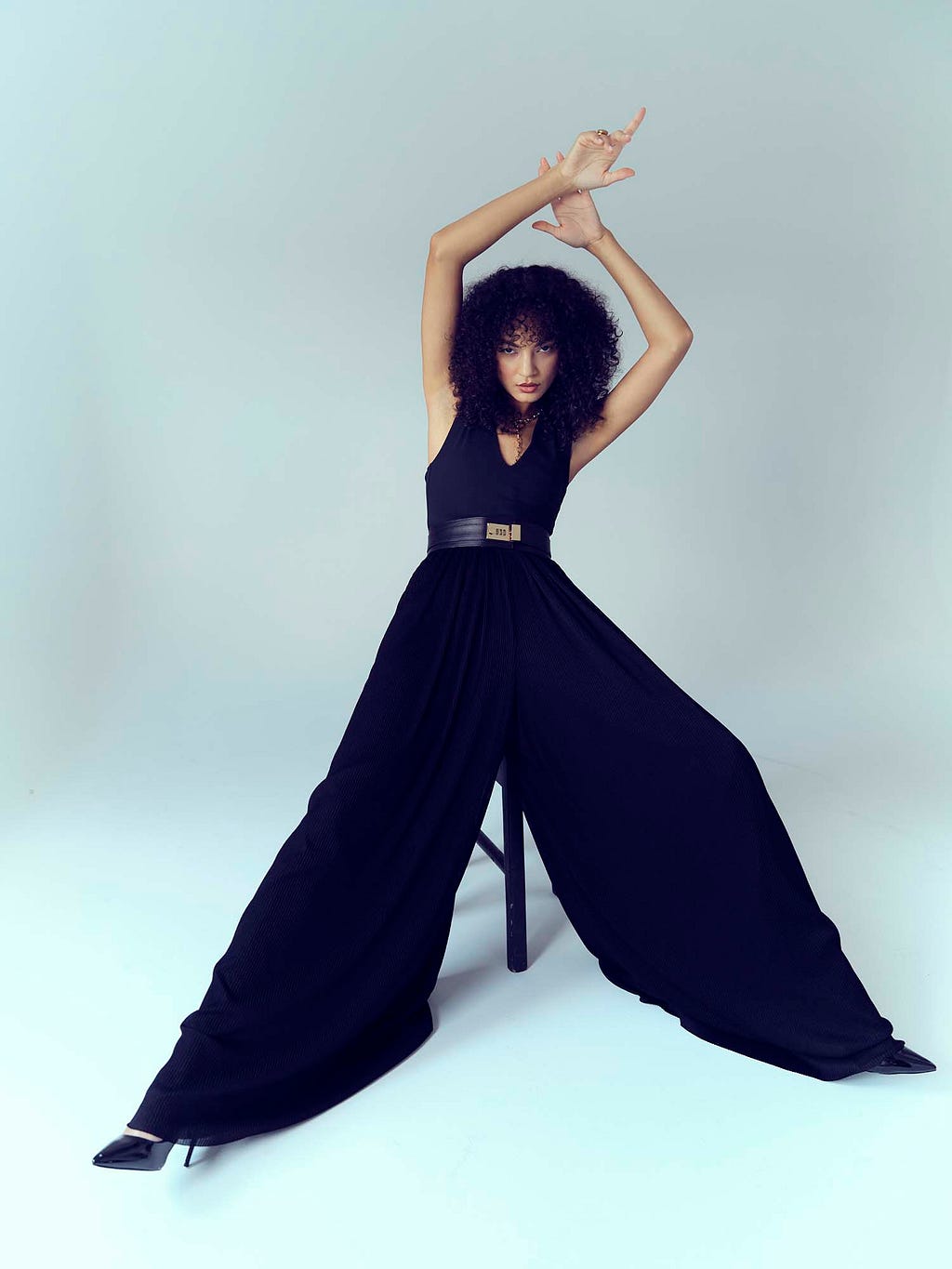 Model in flowing black trousers sat on a chair against pale blue backdrop