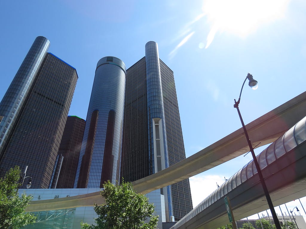 A photograph of the sun shining down over the GM Renaissance Center from the view of right under a People Mover track. One lamppost and two trees also add to its beauty. God bless and I love you!
