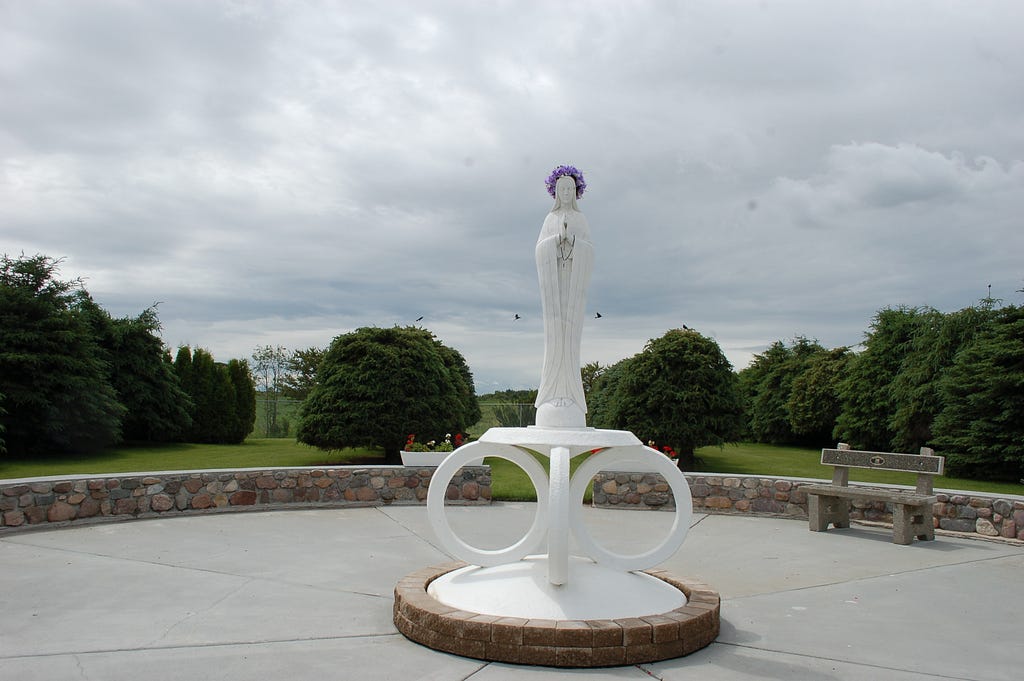 Our Lady of the Highway shrine in Vegreville. A slim white statue of a woman with trees in the background.