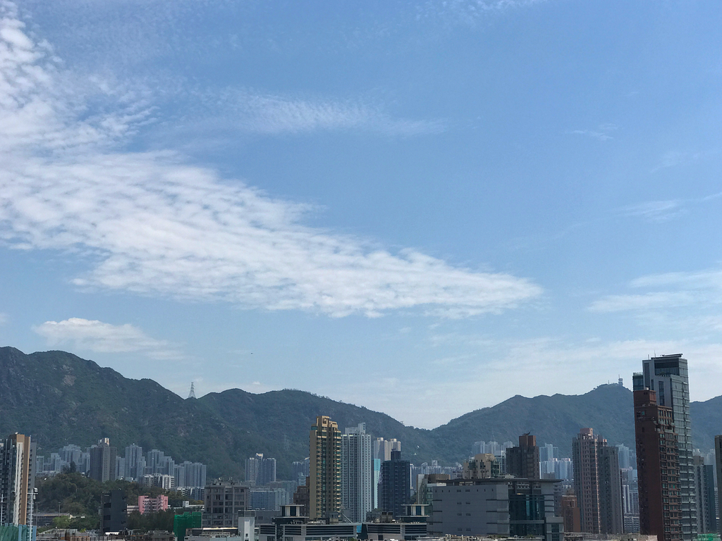 A blue sky with swirling, fluffy clouds over a view of Hong Kong with buildings in front of mountains.