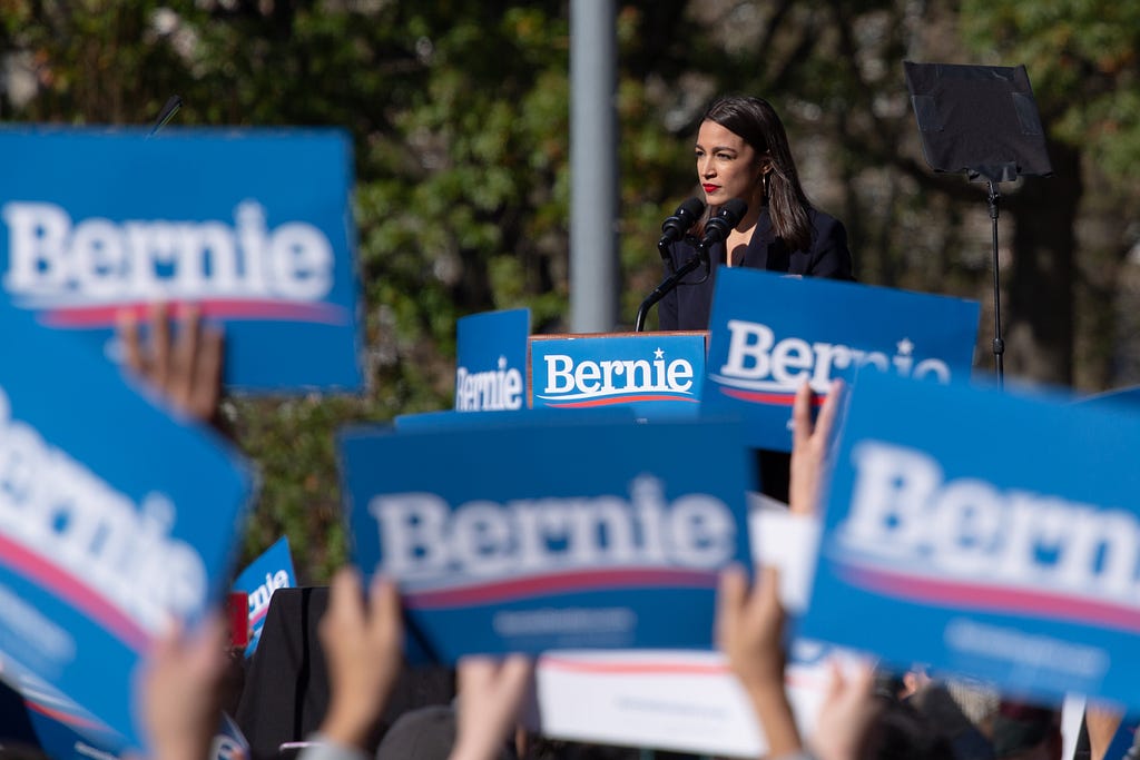 Alexandria Ocasio-Cortez appears confident at a podium with a foreground of Bernie Sanders signs raised from all directions.