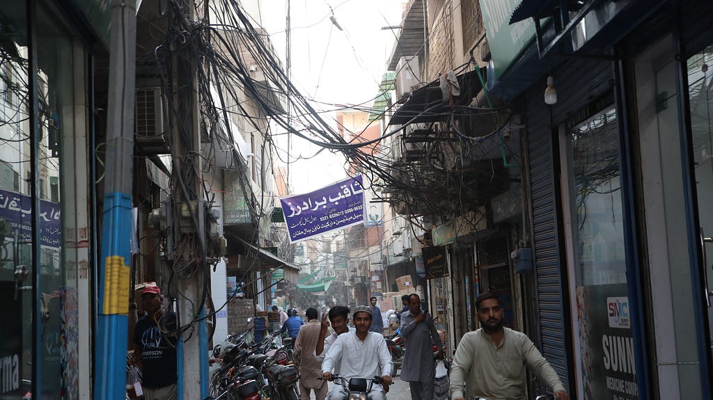 Pedestrians and people riding motorbikes on a narrow city street with a tangle of electrical wires hanging above their heads.