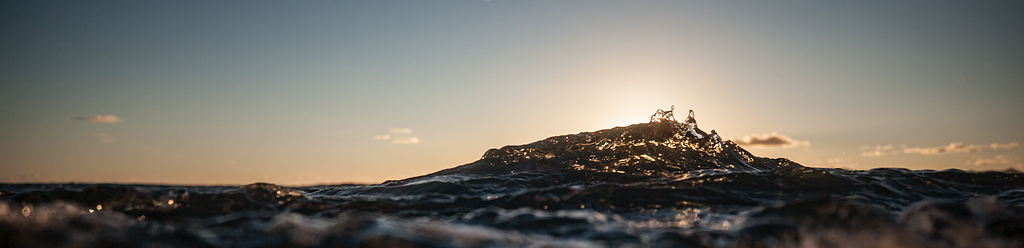 Photograph of a wave at sunset. The peak of the wave is highlighted by the sun.