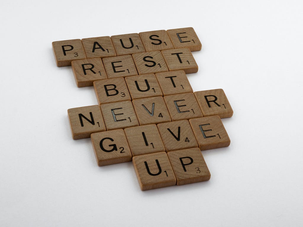 Brown square scrabble tiles spelling out ‘pause, rest, but never give up’ on a white background.
