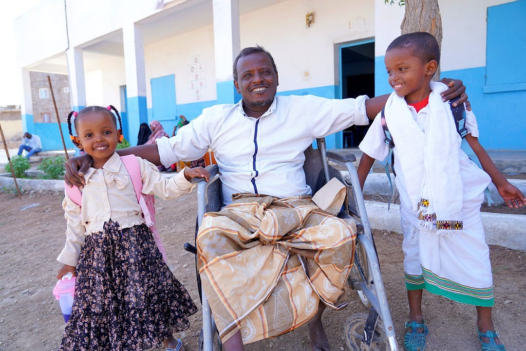 A man in a wheelchair smiles broadly with an arm around his young daughter and nephew outside of the preschool they attend.