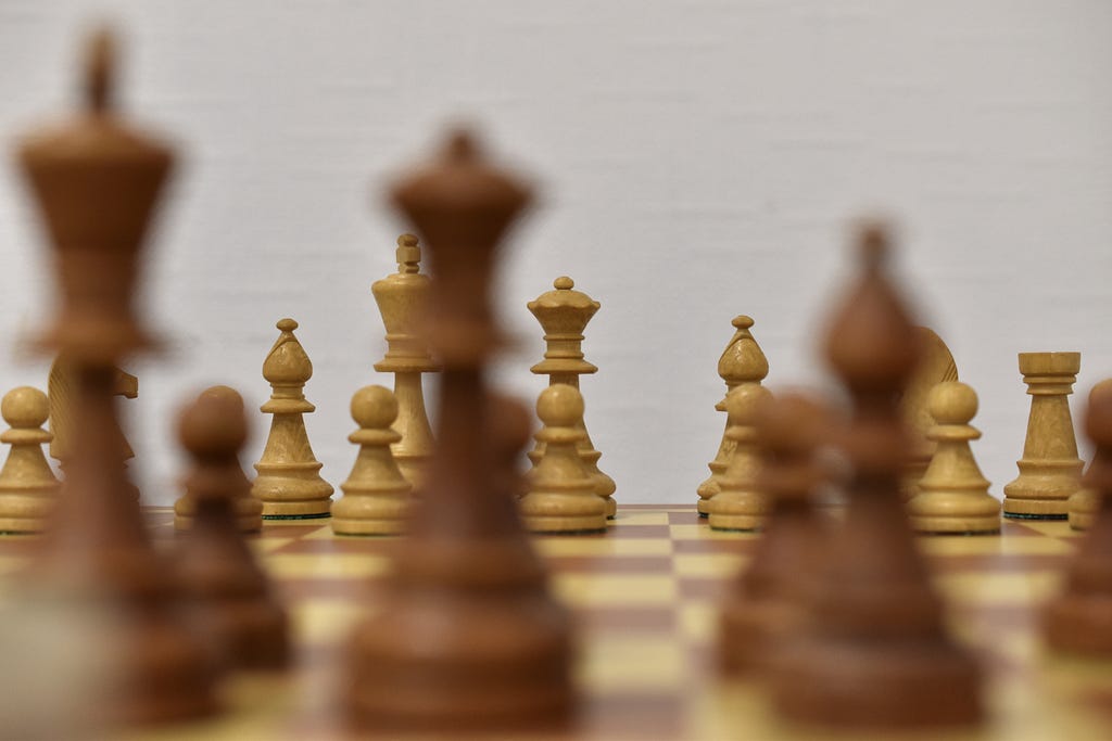 A chess board. White pieces are viewed in the background through blurry black pieces in the foreground.