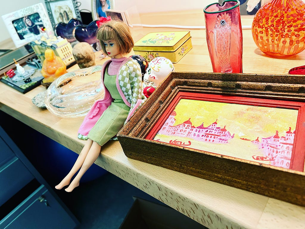 Barbie sits on a desk wearing a green dress next to a pink and yellow picture of venice and assorted vases and pictures.