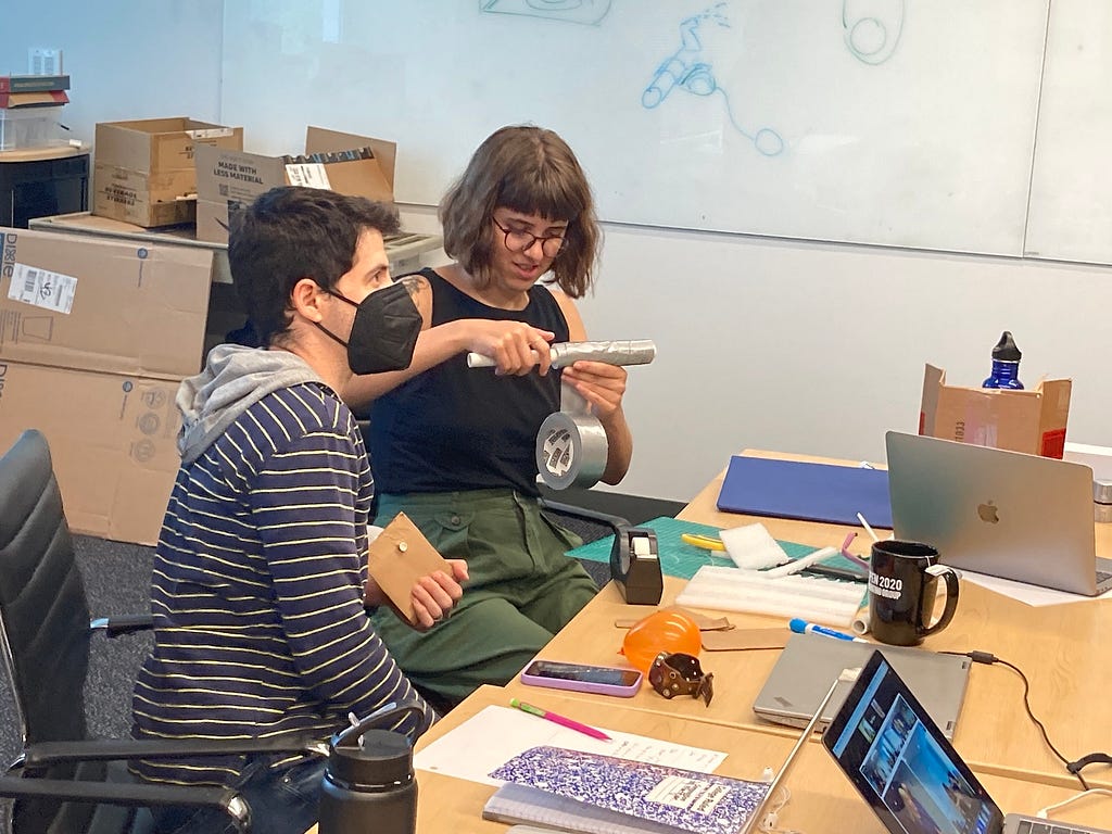 Photo of 2 teachers sitting at a table in an MIT classroom. Teacher on the left is looking at something out of frame, teacher on the right is looking down at the duct tape in her hands. Craft supplies and laptops are strewn on the table in front of them.