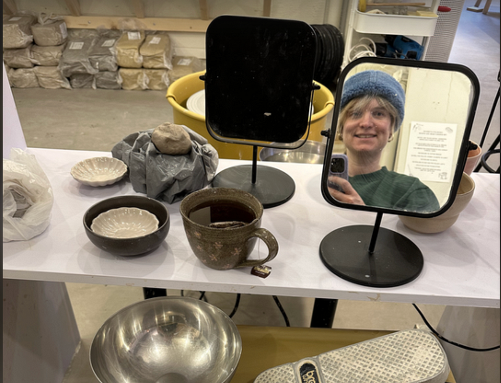 A table of ceramics with a small mirror, reflected in the mirror is Nicole Michaelis wearing a blue beanie hat
