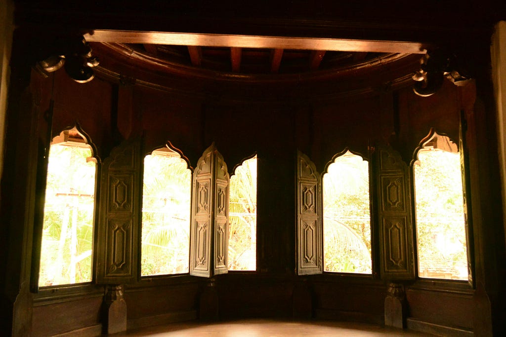 A photograph of a wall full of arabesque-scroll design windows. Each of the four windows has shutters. The room is in a 17th Century-style. Light is streaming in through the windows.