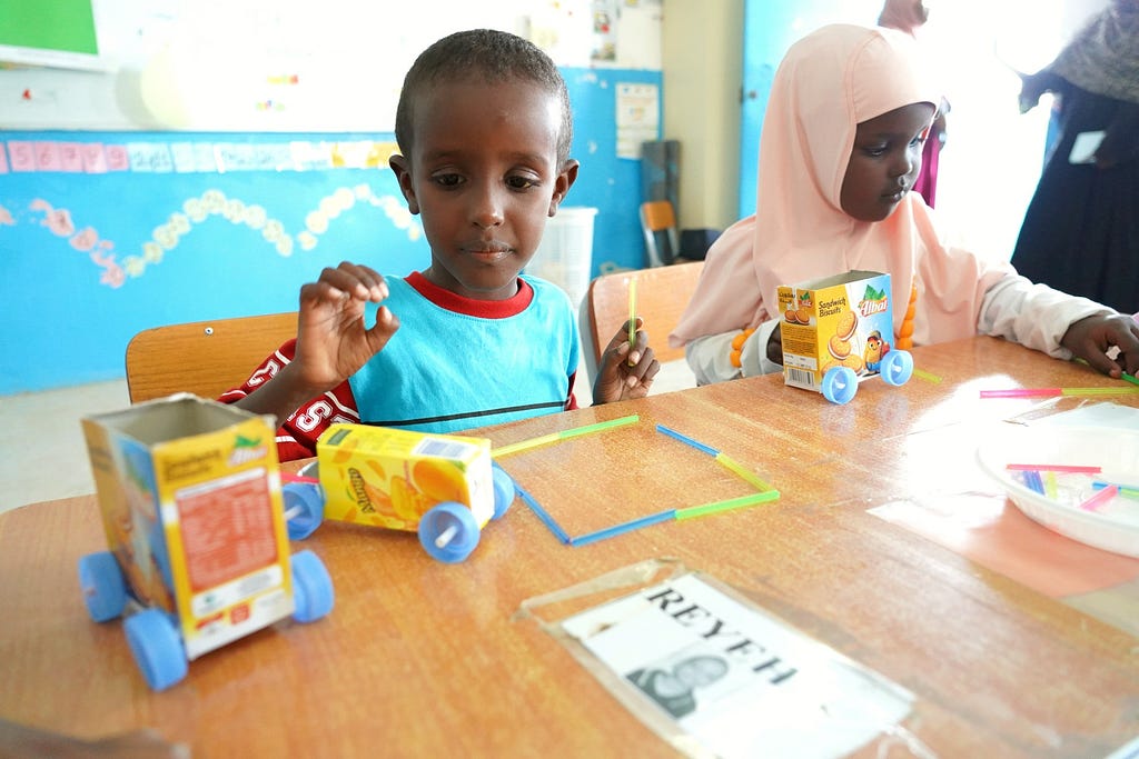 A boy and girl sit at their work stations at school with the toy cars they have built with small recycled cardboard boxes, colorful drinking straws, and little blue wheels.