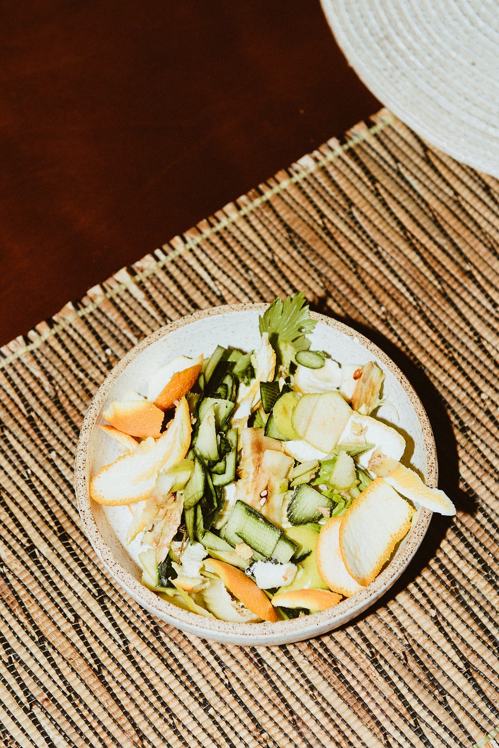 A bowl of food scraps — orange peels, cucumbers, and lettuce — atop a wood placemat.