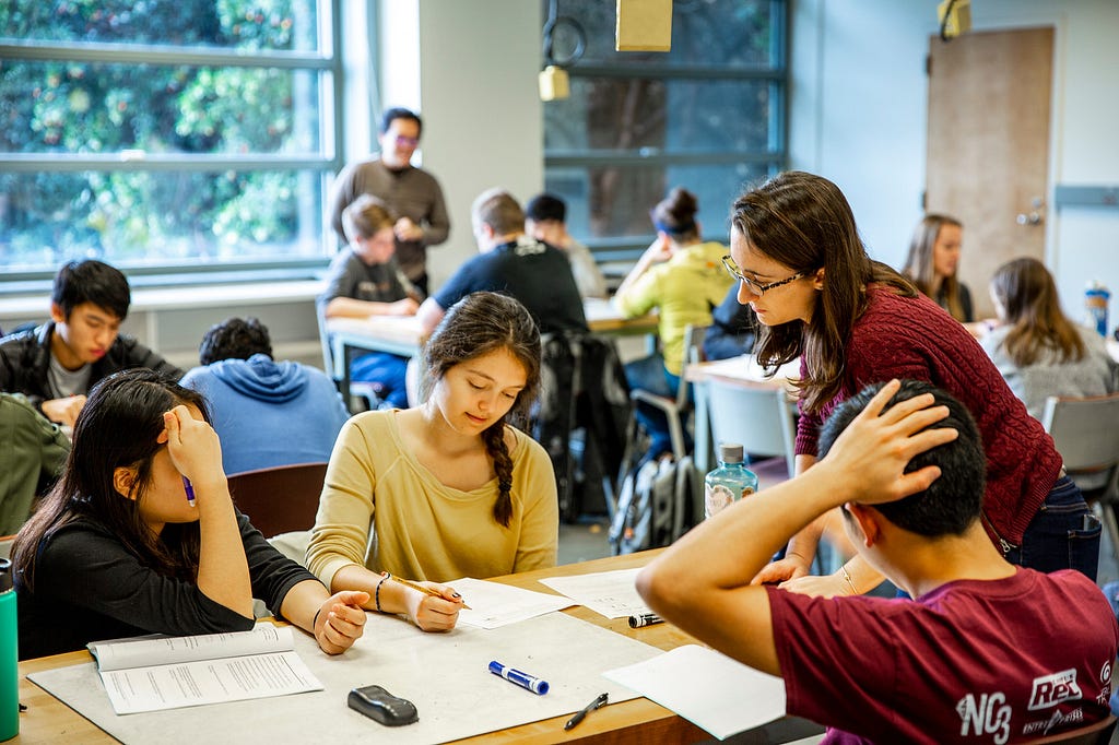 An instructor talking with three students surrounding a table, the students are collaborating on whiteboards and papers.