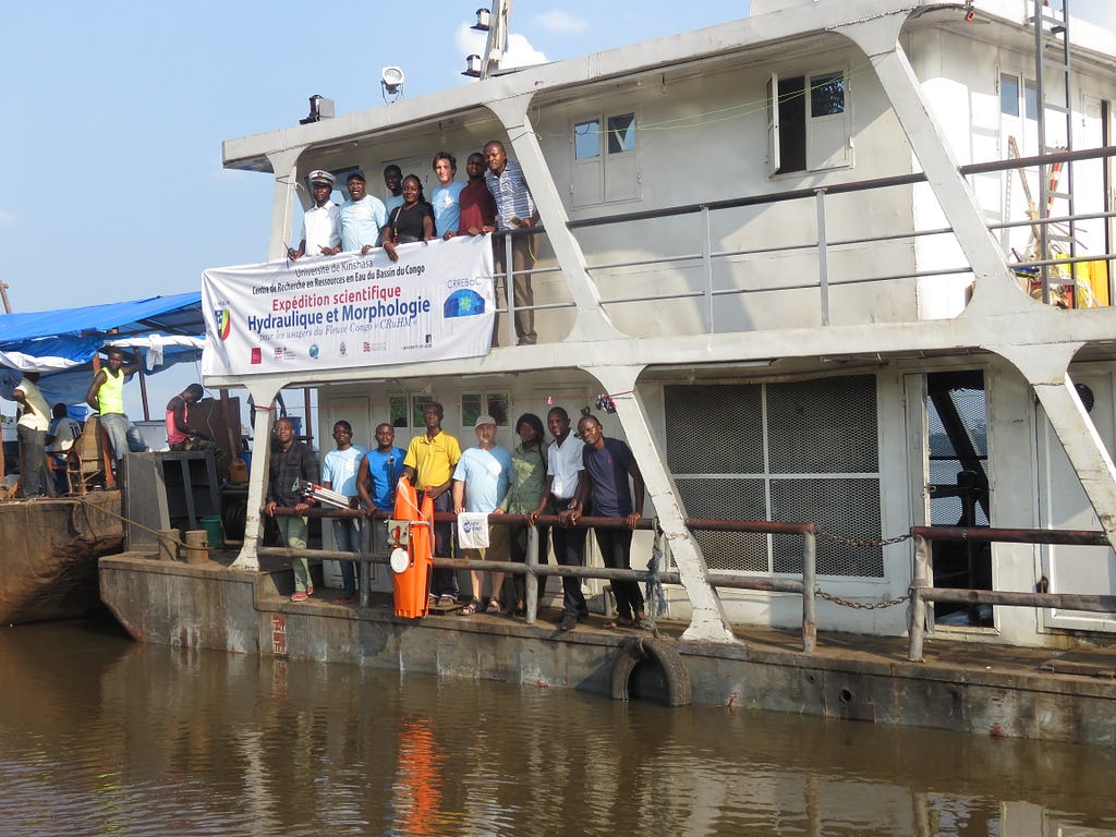 A group of people stood on a large boat sitting on the Congo river. One of them is holding a piece of scientific apparatus