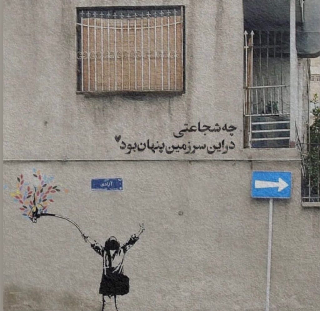 Photograph of the side of a brown cement building in Iran. There is a stencil of a young girl holding her arms up as if in celebration, with a bouquet of flowers in one hand. Above the stencil, graffiti in Farsi script reads ”What courage was hidden in this land.”