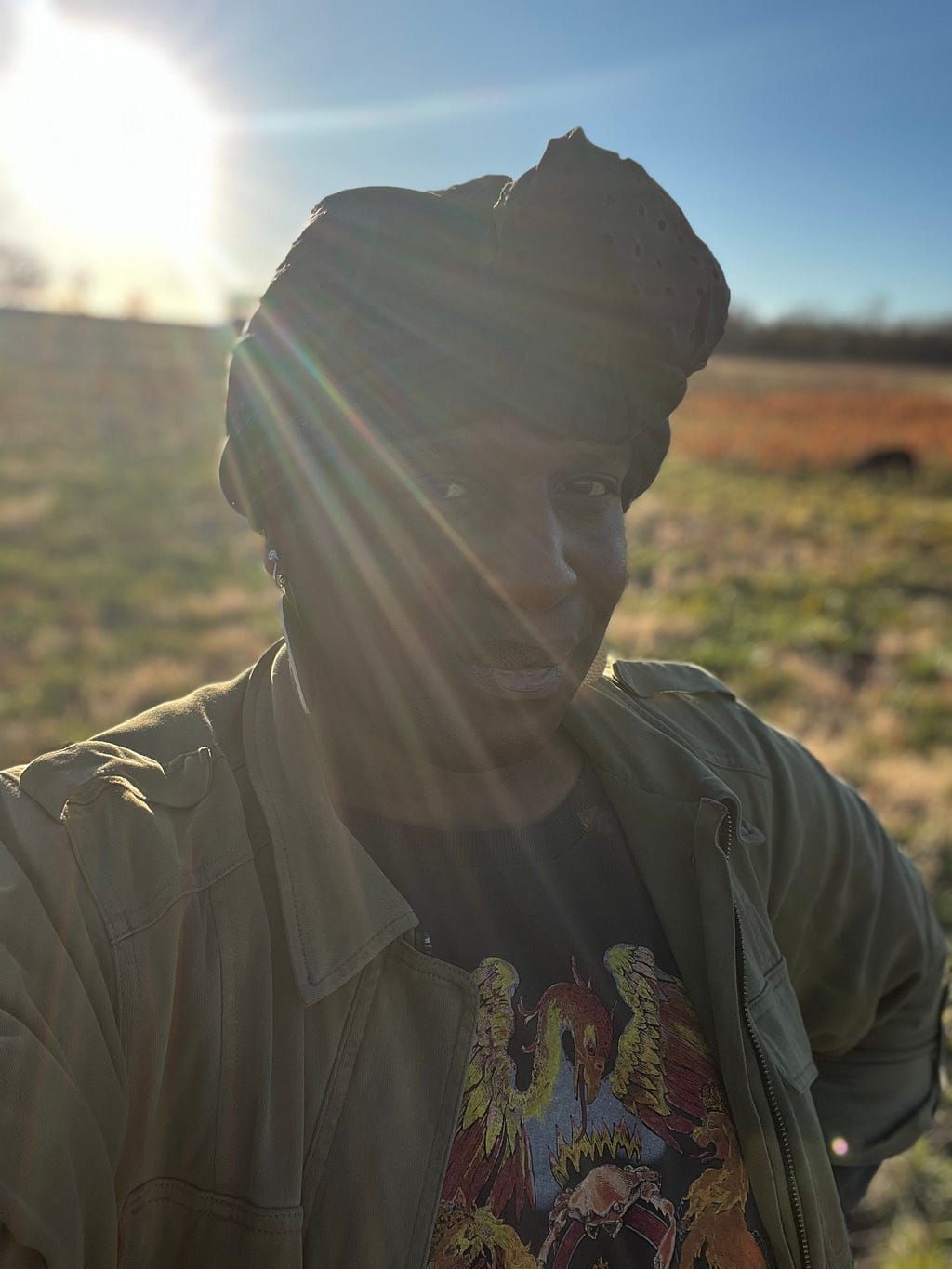 Black trans human standing in a field with the sun on their back. the human has a smirk, as if they know something the reader doesn’t.