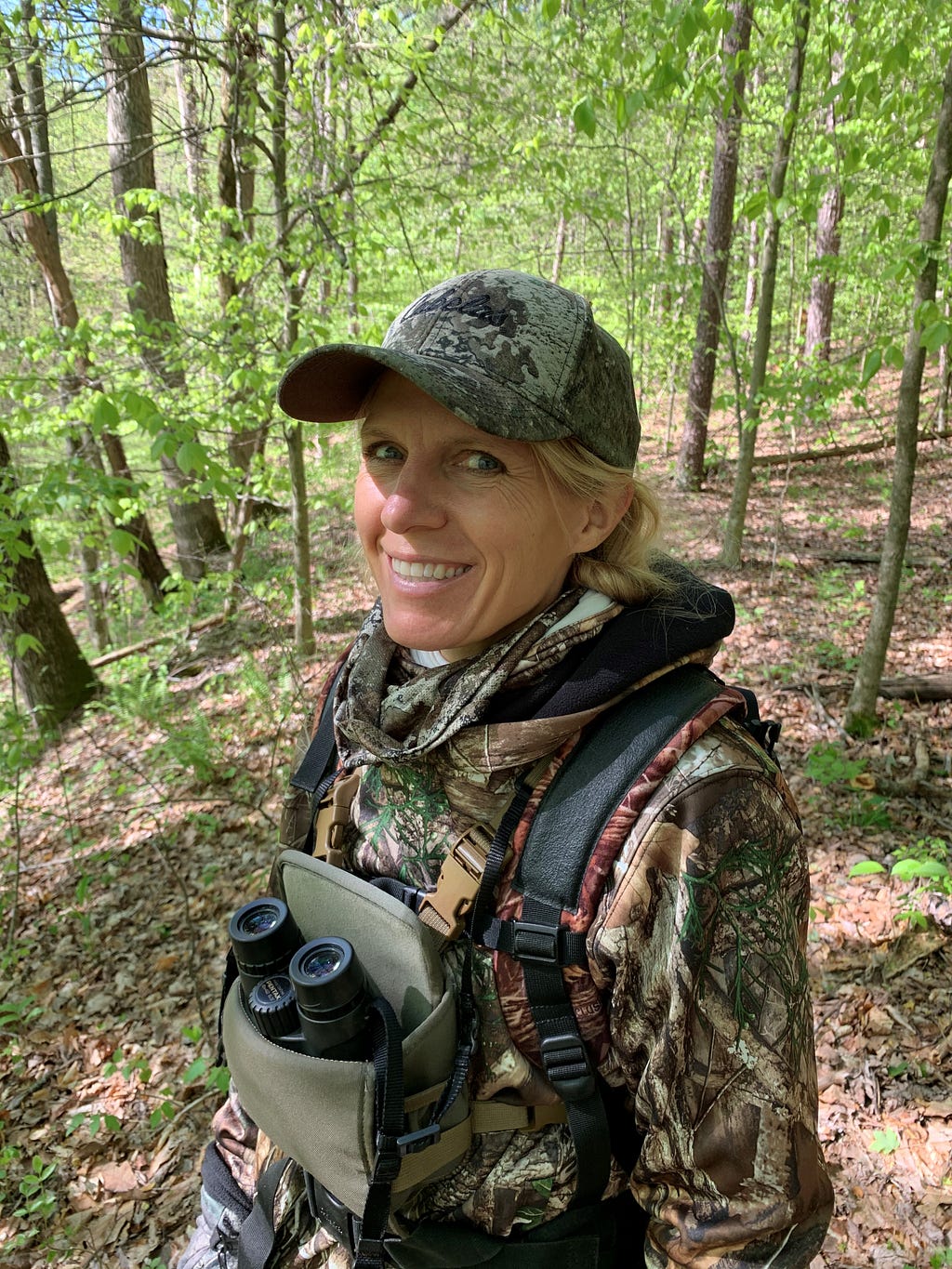 In this profile photo, U.S. Fish and Wildlife Service biologist Joelle Gehring walks in the woods wearing a camouflage jacket and hat and a pair of binoculars in a green case.