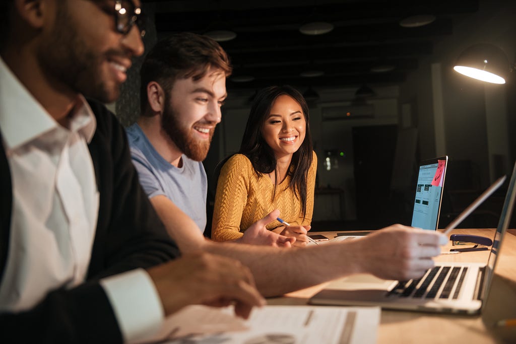 Group of colleagues working late at night in office with laptop and documents. Bearded man pointing to computer with pen.