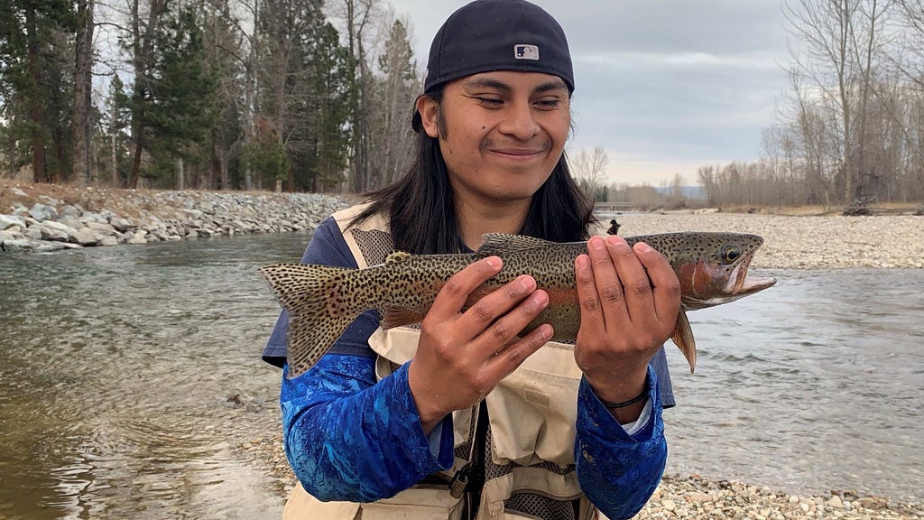 A man holds a fish out in front of him and grins in excitement with a stream and trees in the background