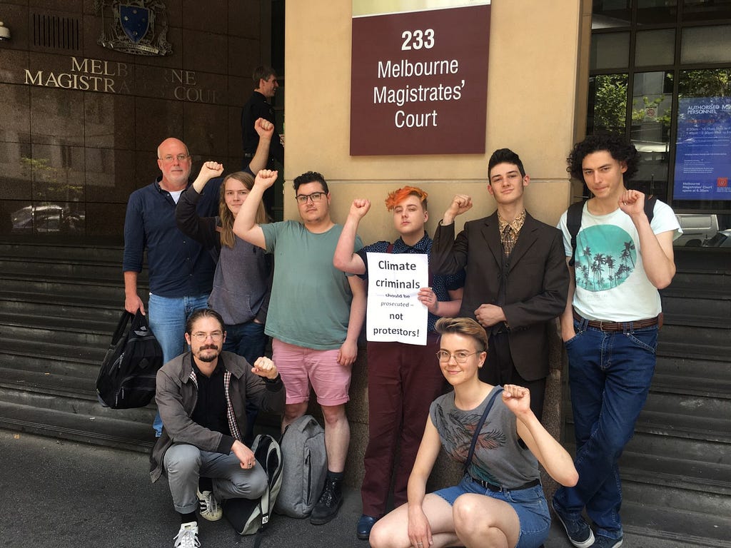 A group of mostly youths, all either standing or crouching by a plaque reading ‘Melbourne Magistrates’ Court’, orange-haired figure in the middle holding up a paper sign reading: ‘Climate criminals should be prosecuted not protesters!’