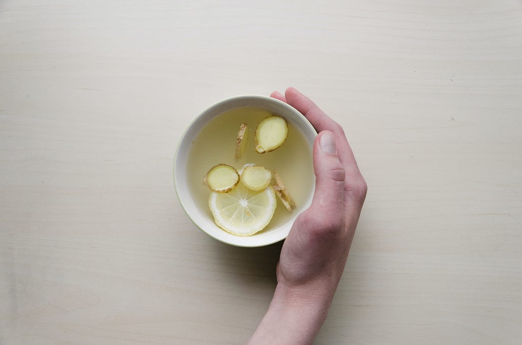 A hand wrapped around a mug of tea with lemon and ginger slices, on a pale wooden table