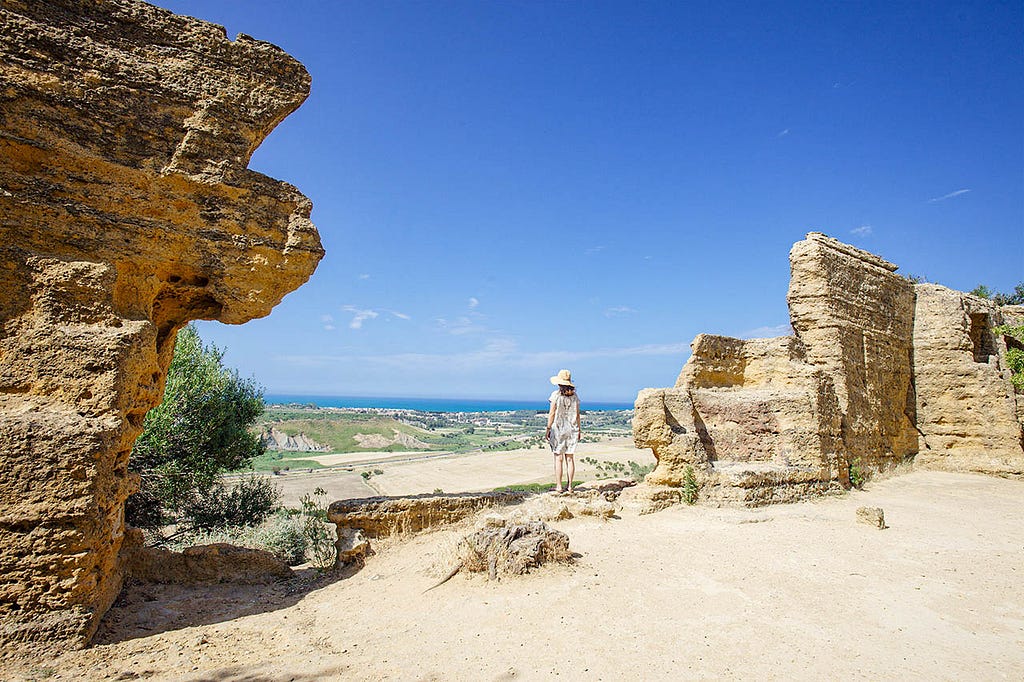 a lady standing in-between ruins looking out at to sea from atop a hill