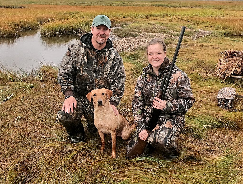 Father, daughter and dog pose on the marsh. Daughter holds a hunting riffle