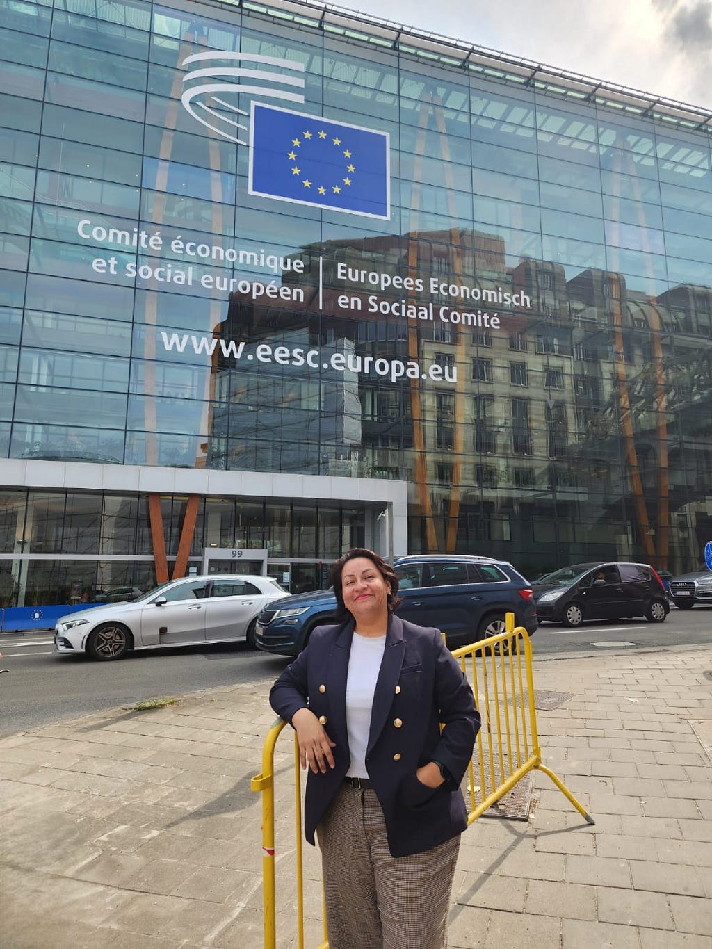 Merary Alvarado in front of the European Social Committee in Brussels, Belgium. Wearing a dark blue blazer, white t-shirt, colorful pants, and short hair.